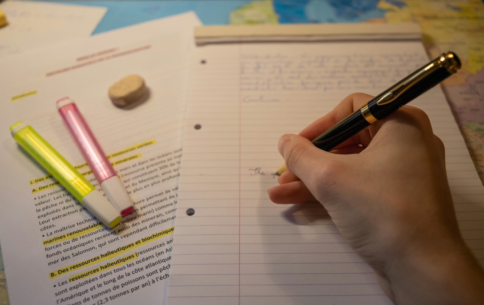 A hand writes notes on lined paper next to a printed reading marked with yellow and pink highlighters