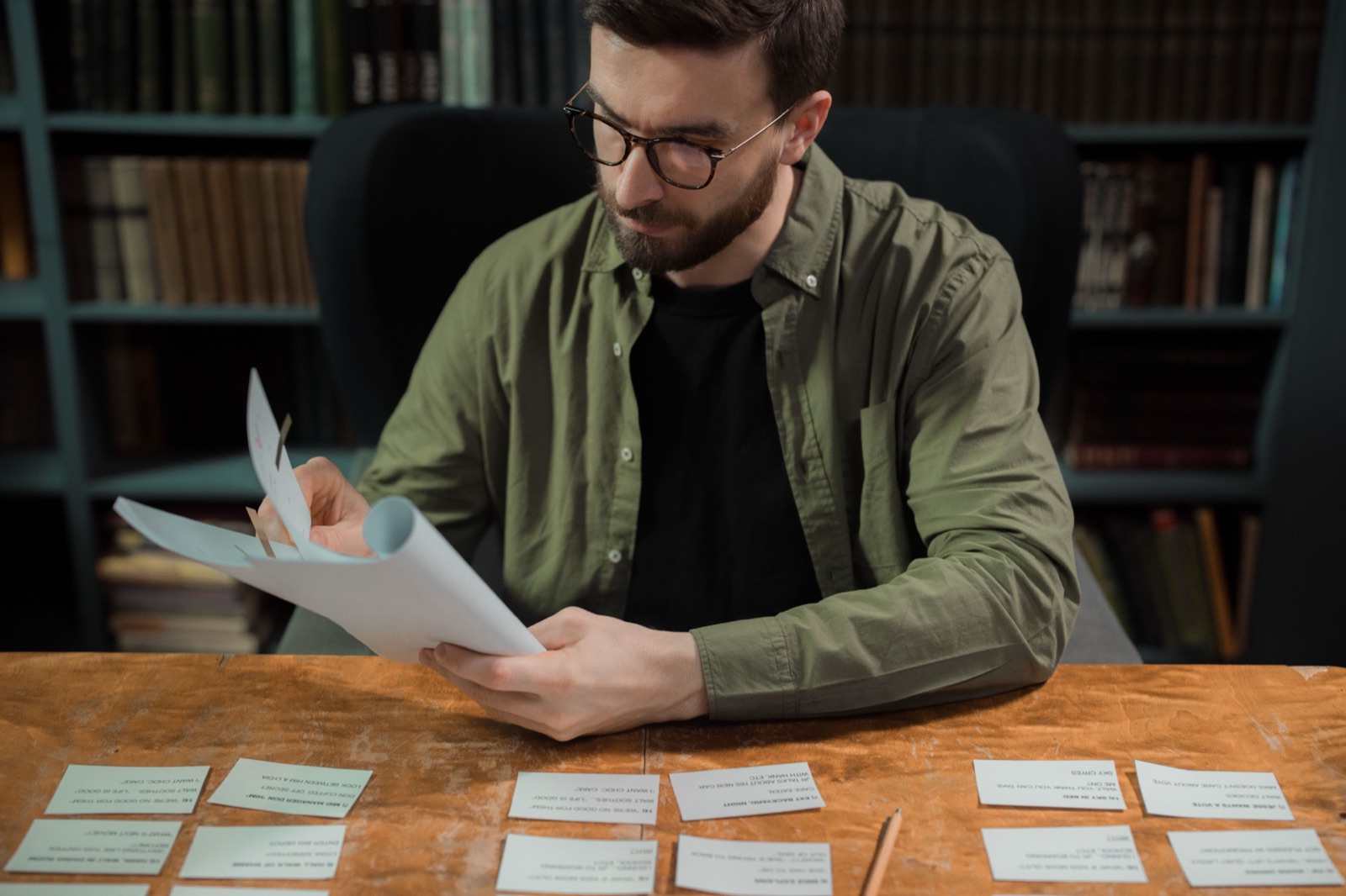 A man in glasses reads through papers at a library table with handwritten index cards laid out in front of him